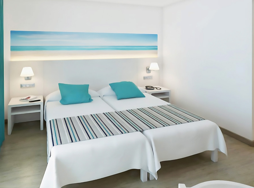 Vista Mar Lateral_Camas Dos camas individuales juntas, con sábanas blancas, cojines azul turquesa y un cabecero decorado con un cuadro de inspiración mediterránea.